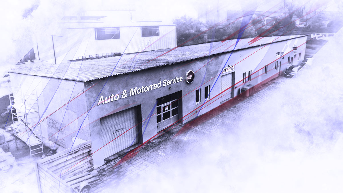 Ein grauer Flachbau mit drei Toren und Türen und acht Fenstern. Als Firmenwerbng ist außen ein großer Schriftzug mit den Worten Auto Motorrad Service angebracht.